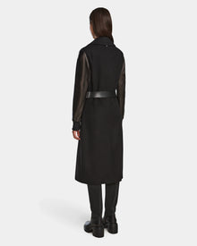 A woman faces away, wearing the Rudsak MARYLIN - 8124520 Black wool coat with leather sleeves, a black belt, black pants, and black boots against a plain white background.