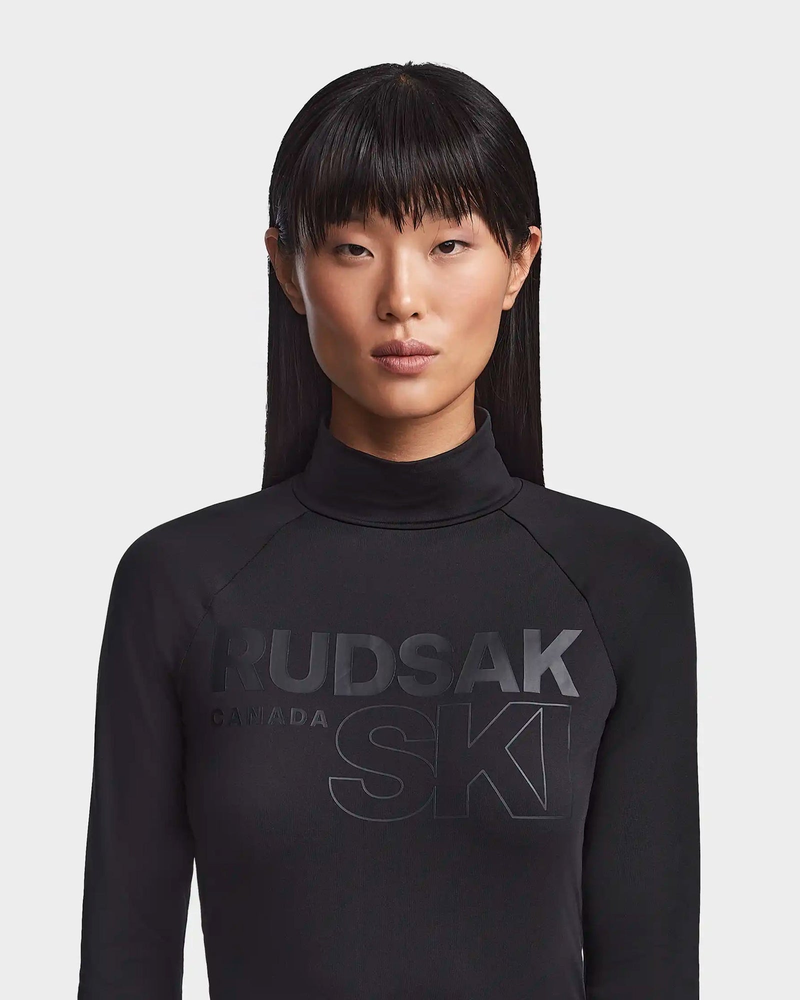 A woman with straight, long dark hair and blunt bangs wears the Rudsak KILTON - 8523533 Black women's fleece-lined jersey turtleneck with bold "RUDSAK CANADA SKI" on the front, elevating any winter wardrobe. She stands against a plain white background.