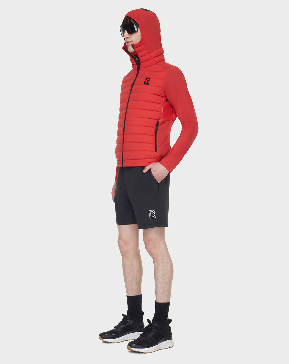 A person wearing a RUDSAK JARRY - 6122007 POPPY RED puffer jacket, black shorts, black socks, black sneakers, and sunglasses stands against a plain white background.