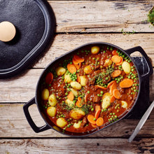 Oval Dutch oven filled with stew, potatoes, carrots, and peas on rustic wooden table