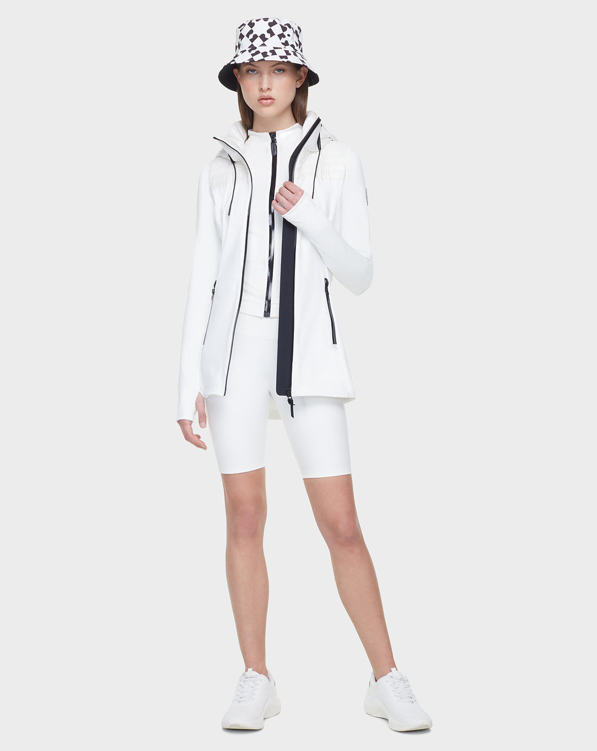 A person stands facing forward in Rudsaks CAMIRA - 8122011 WHITE womens activewear jacket, paired with white cycling shorts, sneakers, and a white bucket hat with a black geometric pattern—perfect for outdoor workouts on a light gray background.